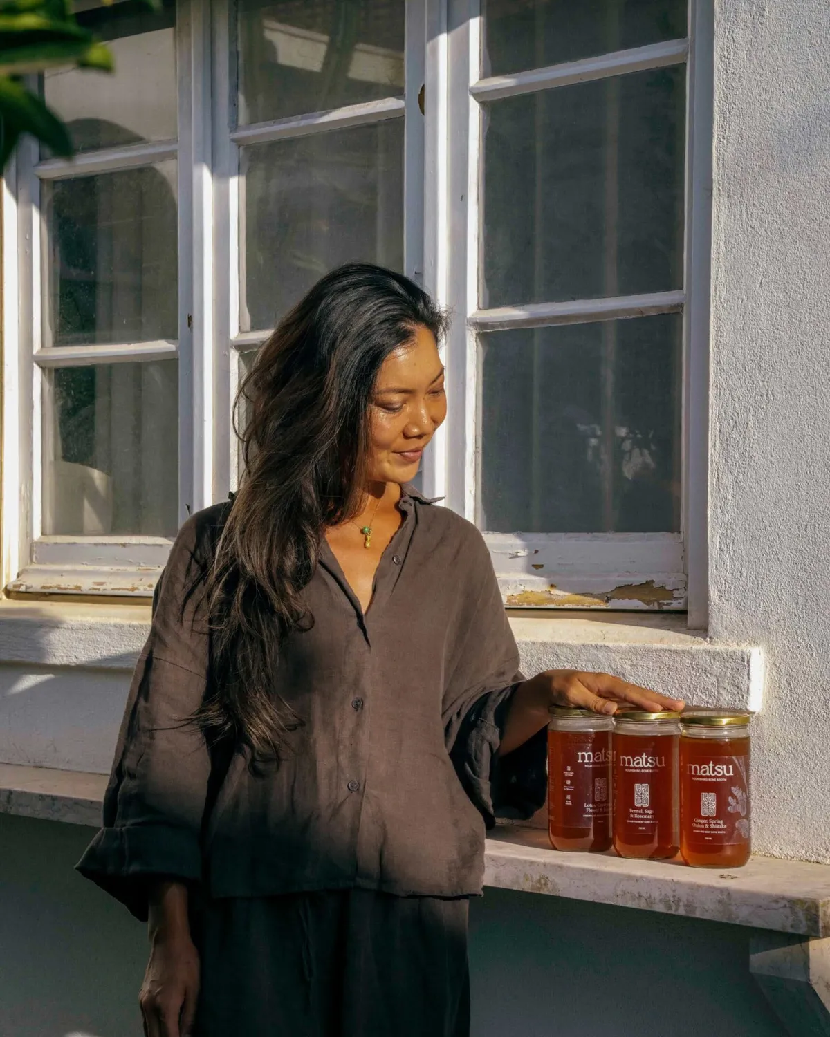 Karen, founder of Matsu, with her jars of slow-simmered bone broth in Ericeira Karen, founder of Matsu, standing in the Ericeira sunlight beside three jars of Matsu bone broth on a windowsill