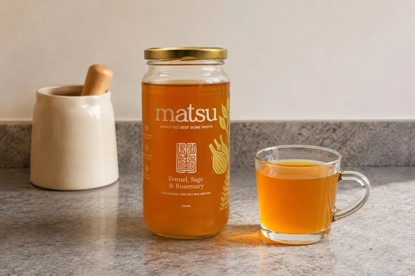 A jar of Matsu Beef, Fennel, Sage & Rosemary bone broth beside a glass cup of golden broth on a stone kitchen counter